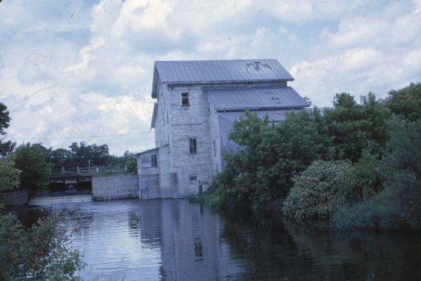 Landscape photograph by Sid Boyum of a fisherman on the bank of Big Spring Creek near a power house and dam. The fisherman to the right blends into the building. 