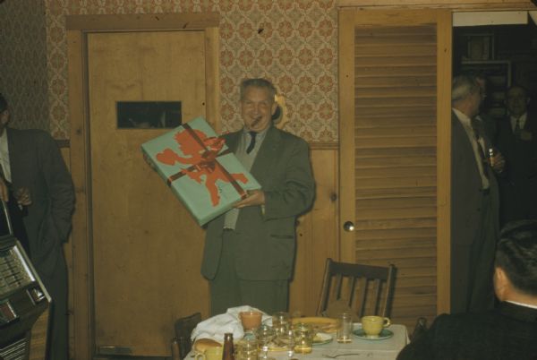 Sid Boyum smiling at the camera, after receiving a gift at a dinner gathering with male colleagues from Gisholt Machine Company. The large, rectangular package is wrapped in mint green colored paper decorated with a red cupid cutout and tied with a satiny red ribbon. Wearing a suit, Sid holds a cigar in his mouth, standing before a table of empty plates, scattered white cloth napkins, beverage glasses and coffee cups, and beer bottles. His colleagues watch him from the sides, by a jukebox, in a doorway and peering through a glass window in a door. They are in a restaurant with wood wainscoting, decorative wall paper, and sconce lighting. 