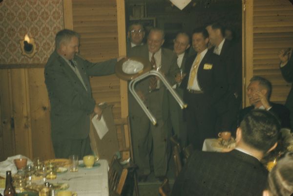 Sid Boyum getting the joke, admiring his gag gift from Gisholt Machine Company co-workers after a dinner gathering. With a cigar in his mouth, he is holding up the gift, a home-made portable "stool" designed with folding metal legs and a wooden toilet seat. His colleagues, sitting at tables and standing in a crowded doorway, are laughing and smiling with him as the are drinking and smoking, too. They are in a restaurant with wood wainscoting, decorative wall paper, and sconce lighting.  