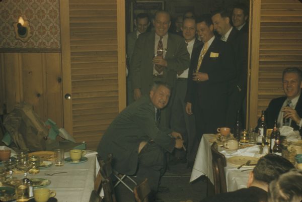 Sid Boyum trying out his gag gift from Gisholt Machine Company co-workers after a dinner gathering. The gift is a home-made portable "stool" designed with folding metal legs and a wooden toilet seat. With cigar in hand, he is seen staring at the camera while seated on the stool with pants down by his knees, thigh visible, and blazer hiding his derriere. His colleagues, sitting at tables and standing in a crowded doorway, are laughing and smiling with him as they are drinking and smoking, too. They are in a restaurant with wood wainscoting, decorative wall paper, and sconce lighting. 