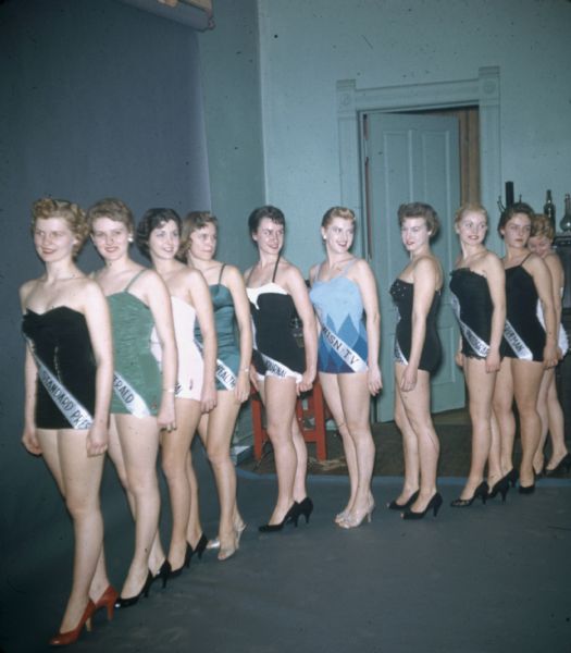 A local beauty pageant for Wisconsin-wide media outlets and community organizations. The line-up of contestants are wearing one-piece bathing suits (with and without straps), kitten heels (peep- and closed-toe), red lipstick and sashes identifying their sponsor ("Miss Eau Claire Press" or "Miss WISN-TV"). Standing at an angle, most of the women have hair pulled back, and some are wearing studded earrings. Looking in several directions, they are standing in a turquoise painted room with a high-ceiling before a blue photographer's backdrop. 