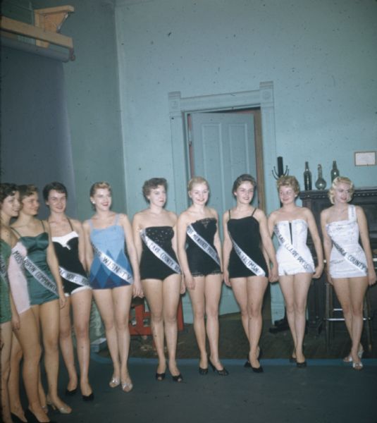 A local beauty pageant for Wisconsin-wide media outlets and community organizations. The line-up of contestants are wearing one-piece bathing suits (with and without straps), kitten heels (peep- and closed-toe), red lipstick and sashes identifying their sponsor ("Miss Eau Claire Press," "Miss WISN-TV," "Miss Lake County Reporter," and "Miss Ripon Commonwealth Press"). Standing to face the camera, most of the women have their hair pulled back, and some are wearing studded earrings. They group is standing in a turquoise painted room with a high-ceiling before a blue photographer's backdrop.