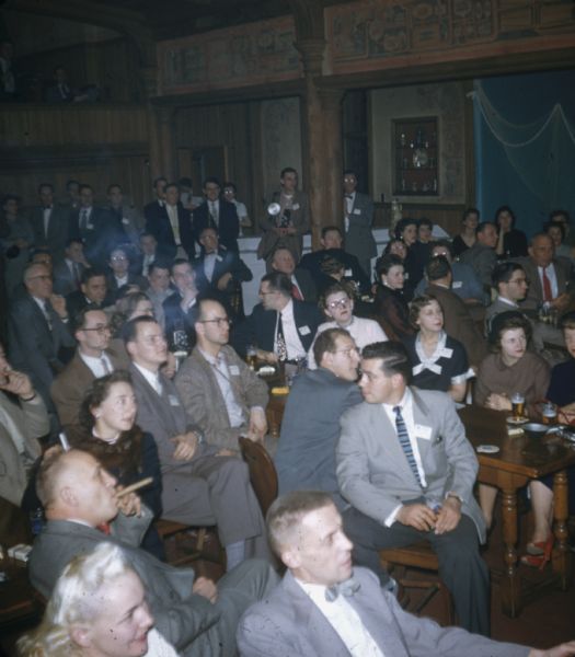 Formally dressed audience members are watching a beauty pageant in a smoky beer hall. The men are wearing suits with ties; the women are wearing dresses and heels, with the occasional fur stole. The audience is sitting together around long tables, and in a balcony (to the back), facing female contestants (out of frame), who are wearing sashes that represent Wisconsin-wide media outlets and community organizations. The audience is drinking pints of Pabst beer, and some men are smoking cigarettes and cigars. Packets of cigarettes are on several tables. A press photographer in the background has a Graflex camera and attached flashbulb. 

The beer hall, known as the Sternewirt taproom in the Pabst Brewing Company, has characteristic German-style thick wooden architectural elements seen in the columns, wall trim, and paneling. There is also a wall niche displaying beer steins arranged around a decorative plate. Brightly colored frescoes painted by the artist, Edgar Miller, in 1943 chronicle brewing history in panels just below the ceiling, and other figural designs decorate the side walls.