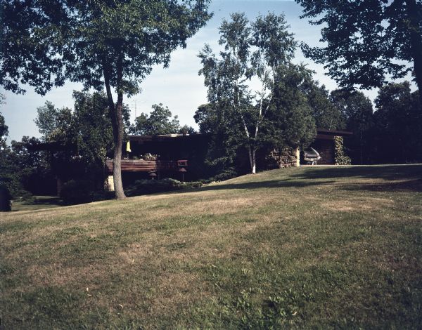 External view across sloping lawn towards a mid-century modern home located by Lake Wisconsin. On the right is a carport framed by stone walls; on the left is an upper and lower level with balcony access, where a closed yellow umbrella stands. There are birdhouses near the balcony, and a tall television antenna is on the roof. The property has a sprawling lawn with trees and garden beds close to the foundation. 

The home was built in 1963 and designed by the architect, Paul A. Thomas III, who taught in the Architecture and City and Regional Planning program at Illinois Institute of Technology. Ken Harley was the original owner from 1963 to 1975. 
