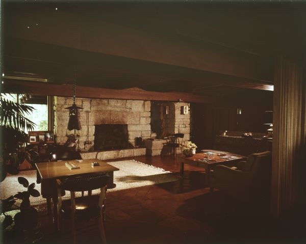 Interior of a mid-century modern home located by Lake Wisconsin. View during daytime of a spacious living room with a large fireplace of cut rock. The wall to the right (obscured) is of board-and-batten construction. Raking light from the right illuminates a low wooden table with magazines and an earthenware ceramic pot holding yellow flowers, and towards the a corner of a large fringed floor rug on a stained and highly polished stone floor. The table on the left has a cribbage board game on it, and two arm chairs set across from each other. A single, Asian-styled lamp is hanging over the table. In the background on the left of the fireplace is a window, showing the roof edge of a lanai. Below the window is a magazine rack. 

The home was built in 1963 and designed by the architect, Paul A. Thomas III, who taught in the Architecture and City and Regional Planning program at Illinois Institute of Technology. Ken Harley was the original owner from 1963 to 1975. 
