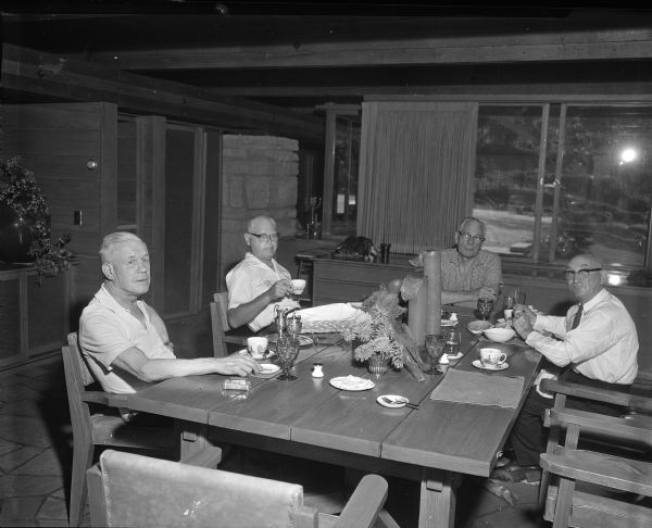 Four men sitting around a long dining table after a casual meal. At the head of the table is Ken Harley, the original home owner who started to build the home in 1960. Three of the men are casually dressed; one is in a necktie and dress shirt. All four are looking at the photographer (Sid Boyum), whose flash bulb is seen reflecting in the back window. The more formally dressed man is eating corn on the cob, and the man across from him is lifting a tea cup. The table includes a vase of freshly cut zinnias, two pillar candles, a small plate with pats of butter, a bread basket, a pack of Pall Mall cigarettes with metal lighter, a silver tea set, and tea cups with matching saucers decorated with pheasants that resemble Elizabethan Staffordshire Bone China. In the bottom right is a dog's paw sticking out from underneath the table near a man's feet.

The dining room shows classic mid-century modern design from ceiling to floor. Large structural cross beams that support the roof of the house can be seen over the table, and dining area walls use horizontal wooden board-and-batten construction. The floor is polished cut stone. 

The home was built in 1963 and designed by the architect, Paul A. Thomas III, who taught in the Architecture and City and Regional Planning program at Illinois Institute of Technology. Ken Harley was the original owner from 1963 to 1975; he started the investment firm Harley-Haydon & Co. with partner Robert Haydon in 1934.