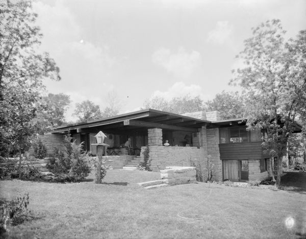 External view across lawn towards the backside of a mid-century modern home. The angle shows the split-level design, marked by wood siding and stone support columns. Two men are sitting in the lanai, or open-sided veranda popular in the 1950s and 1960s. There is a birdhouse on a post in the yard and a garden hose is laying across the mowed lawn. The house was built in 1963.

The home was built in 1963 and designed by the architect, Paul A. Thomas III, who taught in the Architecture and City and Regional Planning program at Illinois Institute of Technology. Ken Harley was the original owner from 1963 to 1975.