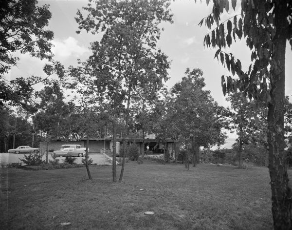 External view across lawn towards the entrance, parking area and lanai of a mid-century modern home by Lake Wisconsin. The lake and a small island are visible through the trees in the distance. The foreground is a long view of the yard with several stands of trees and a couple stumps, cut at ground level. In the middle is the house, where a man in a white shirt is standing under the open-sided varanda, and a parking area with three automobiles — a suburban, a single-door Pontiac coupe and a two-door Chevrolet Impala. 

The home was built in 1963 and designed by the architect, Paul A. Thomas III, who taught in the Architecture and City and Regional Planning program at Illinois Institute of Technology. Ken Harley was the original owner from 1963 to 1975. 