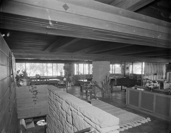 Interior view of a mid-century modern home showing the split-level design and prominent architectural elements of the style. The sprawling open floor plan of the upper level is laterally divided by a stone column and cabinet, and includes a dining area (back left), a living room (to the far right) and a grand piano in the center. A prominent strip of windows that show a stand of trees illuminate the background with natural light that shines off the stained and highly polished stone floor. A stone wall in the foreground demarcates a stairway to the lower level, where a small round table, couch and rug are visible. 

The home was built in 1963 and designed by the architect, Paul A. Thomas III, who taught in the Architecture and City and Regional Planning program at Illinois Institute of Technology. Ken Harley was the original owner from 1963 to 1975.