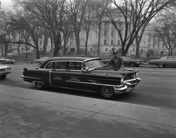 View across sidewalk towards Robert "Bob" Bender standing behind the hood of his shiny 9-passenger black 1956 Cadillac Fleetwood, the flagship taxi of the cab company, which he named "Nellie." He is wearing a chauffeur hat, and his reflection can be seen on the hood. The sign on the rear door advertises the "R.L. Bender Deluxe Cab Limousine Service," and the front door reads "Flat Rate" listing a contact phone number in Madison. The license plate number is E97-070. He is standing on South Carroll Street at his usual taxi stand in front of the Park Hotel at the corner of South Hamilton Street. In the background is the Capitol Building. Bender bought the used Sixty Special model with paired tail fins in 1957, washing it every morning with cold water only, no soap or wax.
