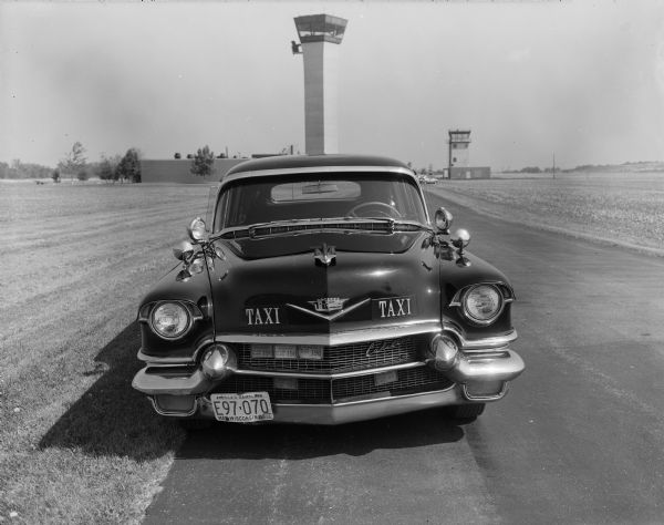 Front end view of Bob Bender's flagship cab named "Nellie," a shiny black 1956 Cadillac Fleetwood, at the Dane County Regional Airport. The straight on angle centers the nose and grill of the car with the air traffic control tower in the background. The license plate number is E97-070 and the text reads: "America's Dairyland Wisconsin Mar '78." Plaques for driver registration "198" and the script Cadillac emblem are attached to the grill; the Cadillac nose logo is broken and half is missing. 