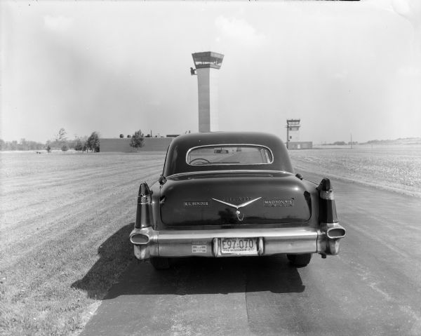 Back end view of Bob Bender's flagship cab named "Nellie," a shiny black 1956 Cadillac Fleetwood, parked at the Dane County Regional Airport. The metal embossed word "Fleetwood" is centered above the chrome chevron, and on either side are embossed business signs that read, "R.L. Bender" and "Madison, Wis." The license plate number is E97-070 and the text reads, "America's Dairyland Wisconsin Mar '78." A small bumpersticker on the left advertises "Don's Transmission Service." The air traffic control tower is centered in the background.