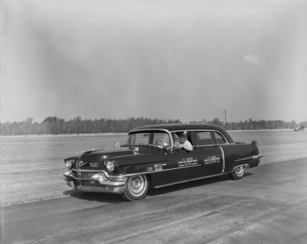 Three-quarter view from front left of Bob Bender sitting in the driver's seat behind the wheel of his cab named "Nellie," a 9-passenger shiny black 1956 Cadillac Fleetwood. Wearing a chauffeur hat, he is leaning slightly out the window, elbow bent on the door. Purchased used in 1957, the limousine was the flagship cab for Bender's taxi company. The signs onthe two side doors read: "R.L. Bender Deluxe Cab Limousine Service." The car is parked on a street along an open field in late summer or early autumn near the Dane County Regional Airport. A person is driving a tractor on the field in the background on the right.