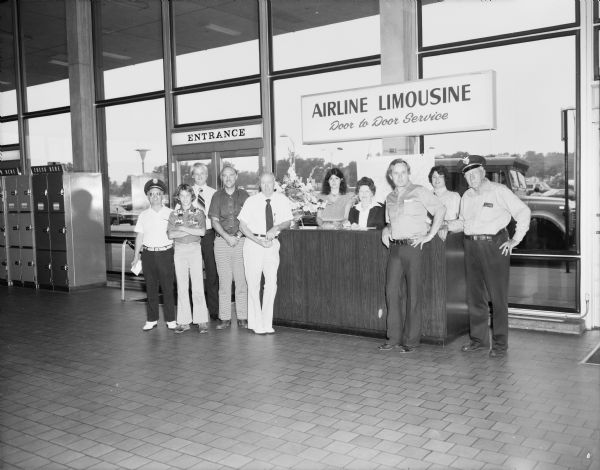 Group portrait of the R.L. Bender Deluxe Cab Limousine Service staff at the Dane County Regional Airport. The owner, Robert "Bob" Bender, is standing on the far right with his hand on his hip and chauffeur hat on his head. The crew is posing by the airport entrance at their service desk under a sign that reads: "Airline Limousine Door to Door Service." Limousines and taxis shuttled passengers between the airport and downtown Madison. On the other side of the entrance are metal lockers under a sign that reads: "Check Here."