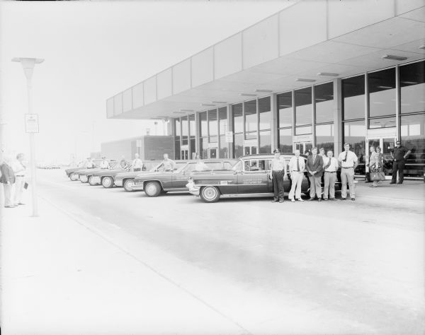 Outdoor group portrait of the R.L. Bender Deluxe Cab Limousine Service crew and taxi fleet in front of Dane County Regional Airport. Robert "Bob" Bender (the owner) is standing in the background to the left of the group in front of "Nellie," his shiny, black, 1956 Cadillac Fleetwood. He is wearing a chauffeur hat. Drivers are standing beside their more modern taxis, that are all angled in a row on a diagonal. Bender's Airline Limousine Service operated sixteen cabs, and all but two of them were Cadillacs, shuttling passengers between the airport and downtown Madison. Two men are standing on the sidewalk on the far left, and other people are standing on the sidewalk in front of the airport entrance on the far right.