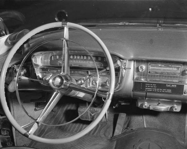 Interior view of the steering wheel, dash, and foot pedals of Bob Bender's deluxe limousine cab named "Nellie," a black 1956 Cadillac Fleetwood. Flat rate fees are written on the dash above the radio: "$10.00 per hour or 50 cents per mile" and a "25 cent toll gate charge at airport to be paid by the customer" per ordnance #2195. The odometer reads: "99999.960." 

He and Nellie reportedly racked up 250 miles a day on average since 1957, when he purchased the car used; at that time it had 40,000 miles on it. In May 1981, Nellie had 993,000 miles; and in August 1982, it was about 1,003,000. Bender owned the company, "R.L. Bender Deluxe Cab Limousine Service," that ran passengers with an additional fleet of later model taxis between the Dane County Regional Airport and Madison downtown.