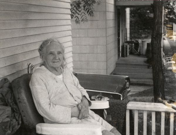 Emma Marie Boyum (nee Schroeder), mother of Sid Boyum, sitting in a large chair on the front porch of her home at 237 Waubesa Street on the east side of Madison. The top of a small garden rake is jutting out from behind her, and a sumac tree branch is hanging over a car parked on a driveway between her home and the neighbor's. The sidewalk up the street towards Atwood Avenue is visible in the far right of the photograph. 

Census records show that Emma began living at this residence with her younger brother, Edward, and her two youngest children (Sid and Bernice) since at least 1929; she maintained residency until her death, when Sid inherited the home. She died on October 27, 1974, at the age of 94. 