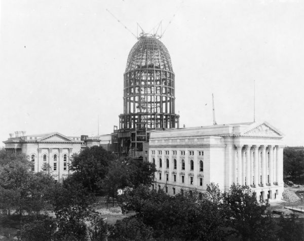 Elevated view over trees towards the Wisconsin State Capitol during construction of the dome. The wing in the foreground is the new west wing. The old north wing is on the left.