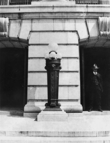 Lamppost outside of the Wisconsin State Capitol. In the entrance cove of the building on the right is a man wearing a suit, standing partially in shadow. 