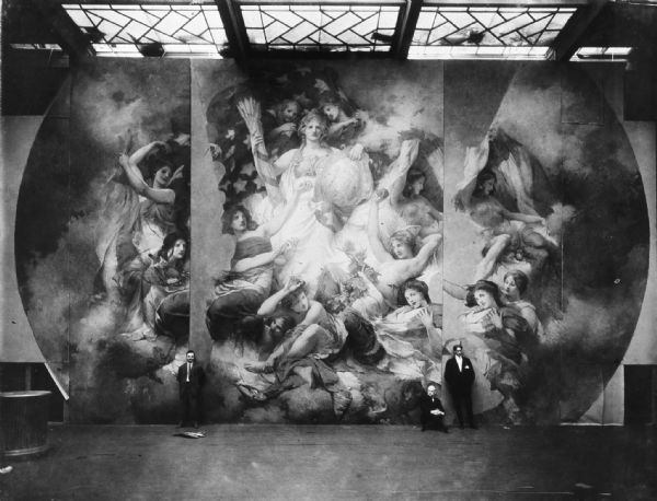 Three men are posing in front of the "Resources of Wisconsin" mural.

The only room in New York City with proper lighting and space large enough for Edwin Blashfield, the artist, and helpers to work on a 34 foot painting was the Vanderbilt Gallery in the Fine Arts building.

When the mural was finished, it was cut, with the paint still fresh, Grandmother said, into sections. Then they were rolled up, placed in boxes and sent to Wisconsin. Here the pieces were spread out and allowed to dry. Then they were re-rolled, put in heavy burlap bags, re-crated and stored in the corridors of the capitol until they could be installed.
 
When that time came, a scaffold was erected, and Blashfield’s assistants came to Wisconsin to supervise the installation, touch up the seams and put a coat of wax on the decoration.