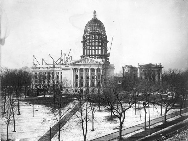 Elevated view of the Fourth Capitol and dome under construction, while the north wing of the third Wisconsin State Capitol remains standing on the right. This view shows the East Pediment, called "Liberty Supported by the Law," by the sculptor Karl Bitter.