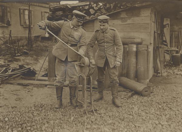Two German soldiers using the services of a field carpentry detachment to sharpen a saber using a hand cranked sharpening wheel outdoors next to a shed.