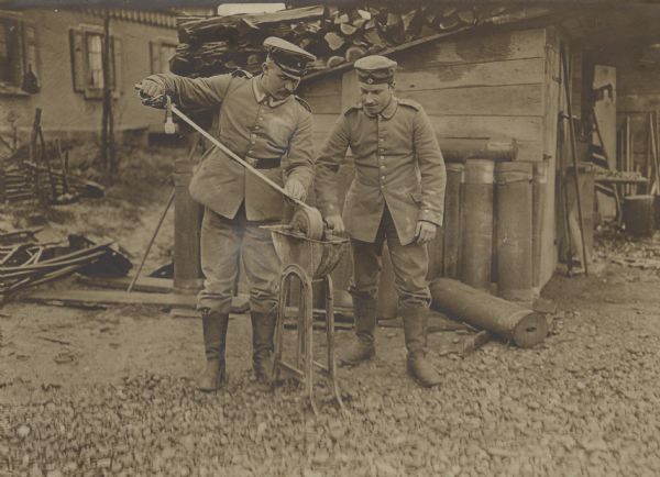 Two German soldiers using the services of a field carpentry detachment to sharpen a saber using a hand cranked sharpening wheel outdoors next to a shed. 