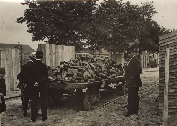 Men and children are standing around a cart which has a large pile of footwear on it for troops on the battlefield.