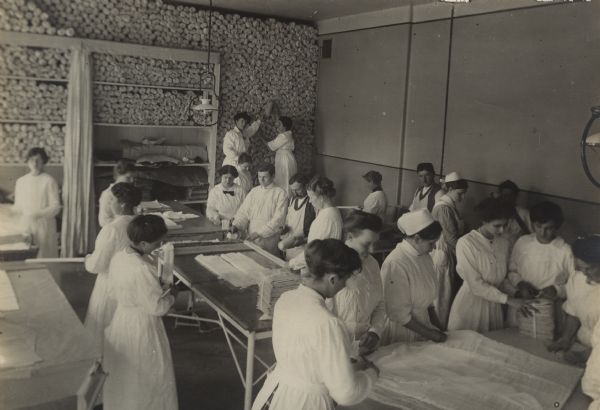Slightly elevated view of nurses, including men and women, packaging bandages for wounded soldiers at a medical depot in Berlin. The group is preparing the bandages on the tables in the foreground, and in the background other nurses are stacking them from floor to ceiling against the far wall.