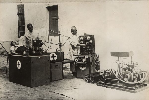 Two men are are standing outdoors and operating a Rotax X-Ray machine on the leg of a man lying on top of a box. Behind them is the side of a building.