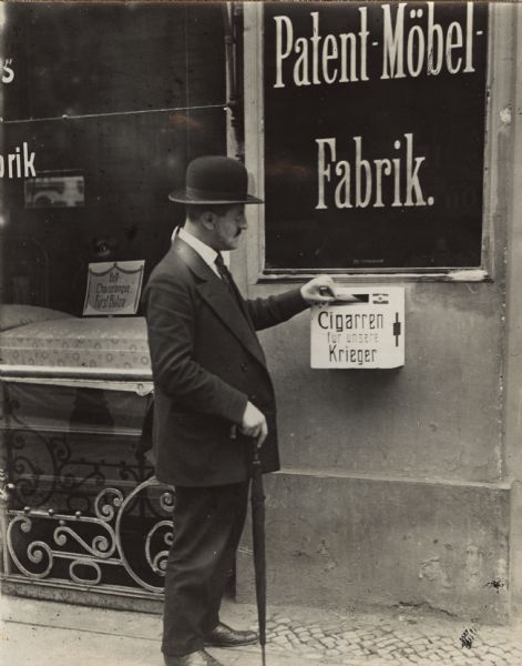 A man is standing on a sidewalk in front of a storefront and donating a cigar to a collection box. A sign on the collection box reads: "Cigarren für unsere Krieger," (Cigars for our Warriors). A sign above the box for the store reads: "Patent-Möbel-Fabrik," (Patent-Furniture-Factory). There is furniture on display in the show window on the left.