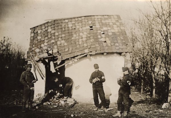 Three soldiers are standing in front of the destroyed house, and another soldier is bending over a pile of rubble in the doorway under the partially collapsed roof.