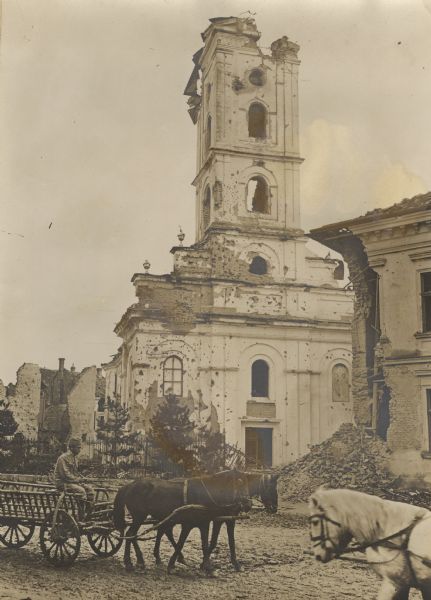 A man is driving a team of two horses pulling a wagon on a street in front of a ruined church. Rubble is piled in front of another building on the right.