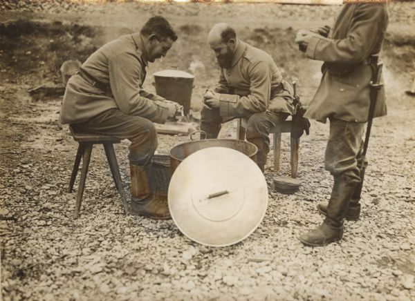 Two soldiers are sitting on stools outdoors peeling potatoes near a large pot, with a cover resting in front on the ground. Another man is standing on the right.