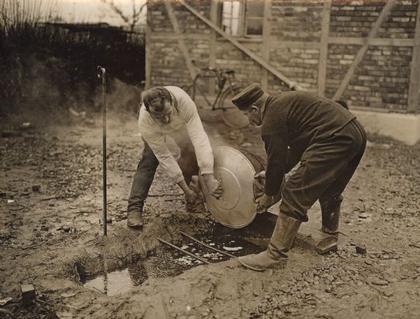 Two men in a muddy yard are bending over and holding onto a large pot while draining water out of it into a trench in the ground. Behind them is a bicycle parked next to a building.