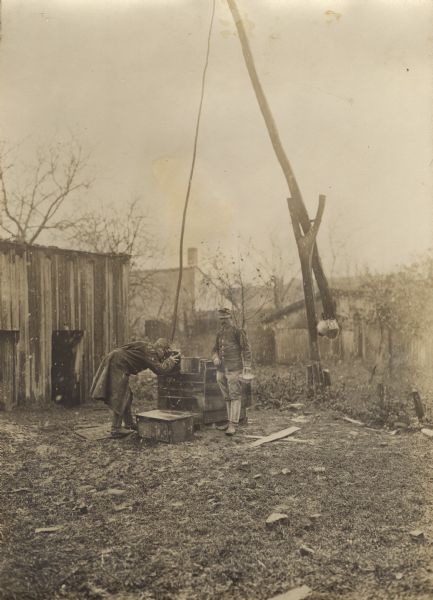 A soldier is using a draw well in a yard and is drinking water from the bucket. Another man is standing nearby. It appears to be raining or snowing.