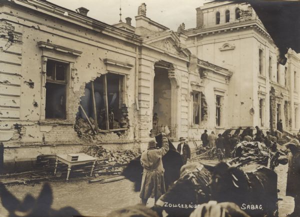 Slightly elevated view of soldiers and civilians near the damaged Customs Building. Broken furniture and rubble is piled in front of the building. There are also people inside the building looking out of the broken windows. In the foreground are horses attached to a wagon.