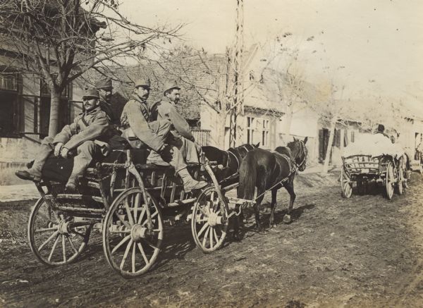 Soldiers are riding in a wagon on their way to the Front. Another wagon is in front of them on the right. There are buildings along a sidewalk in the background.