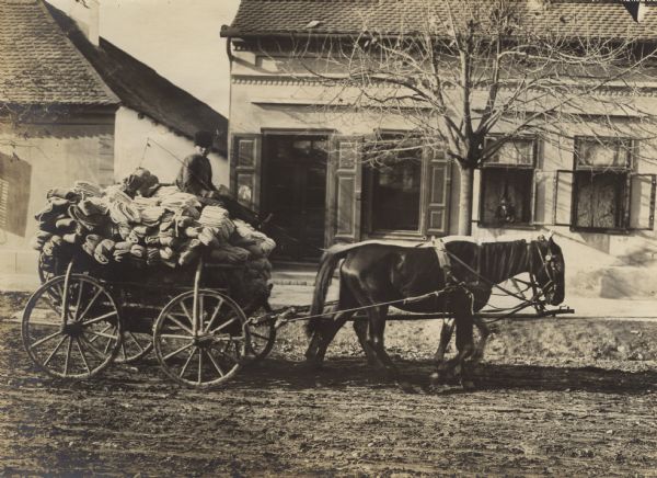 View from side of street towards a boy sitting on top of a pile of supplies driving a horse-drawn wagon heading to the Front. A person is watching from the window of a building in the background.
