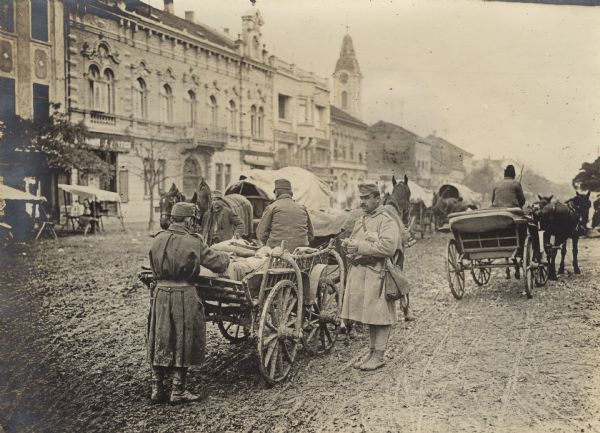 Men are standing on a city street near wagons carrying supplies to the front. 
