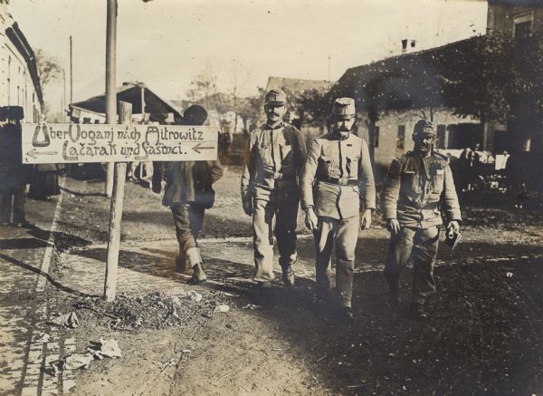 Austrian soldiers are passing a directional sign in the City of Ruma in the region of Syrmien in Serbia. The sign directs traffic to the towns of Vogani, Mitrowitz, Lacarak, and Sasinici.