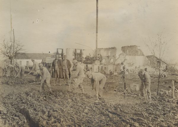 Austrian soldiers are using the stones of a destroyed factory to repair and improve a roadbed. The walls of the destroyed factory are in the background.