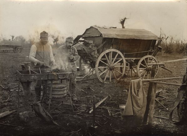 Two soldiers with a small forge during World War I. One of the men is smoking a pipe. There is a wagon in the background.