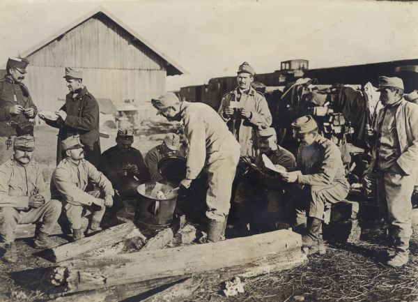 Soldiers relaxing and eating around a man cooking over an outdoor fire. 