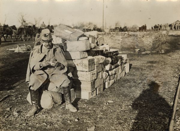 A soldier is sitting on sacks near a pile of boxes while guarding a field post in Serbia during World War I. Horses and men are in the background.