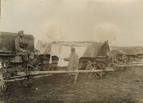 Three Austrian soldiers are sitting and standing around wagons loaded with pontoons that will be used to bridge the Save (Sava) River during World War I before the invasion of Serbia. One of the men is standing near a laundry line.