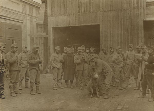 Austrian soldiers are standing around at the train station in Ruma, Syrmien. A man is standing in the center with a sheep, presumably about to be slaughtered for a meal.