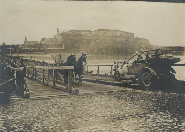 A car and a wagon are crossing the bridge over the Danube in World War I. Pedestrians are using the walkway of the bridge on the left.