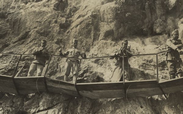 View looking up at Austrian soldiers standing on a wooden walkway with a metal railing attached to the side of a cliff above a lake in South Tyrol.