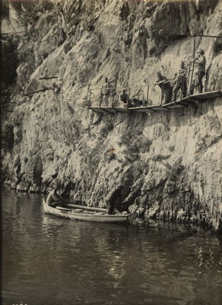 View across water towards Austrian soldiers working on the walkway along the side of a mountain close to the front in South Tyrol. Two men are in a boat at the base of the cliff. Five men and a dog are on a wooden walkway halfway up the cliff.